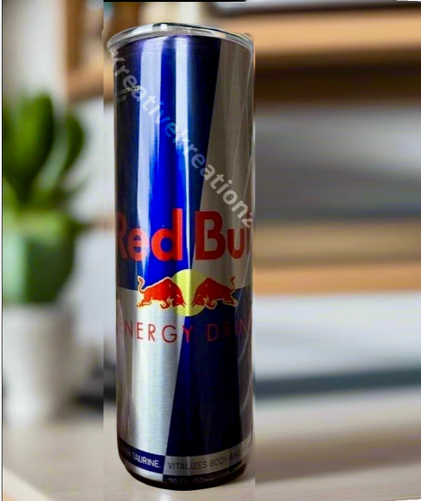 A Red Bull Stainless Steel 20oz Tumbler by Kreative Kreationz stands beside a classic Red Bull energy drink can, its sleek design contrasting with the can's blue and silver colors against a backdrop of blurred shelves and books.