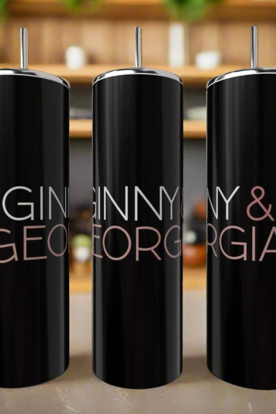 Three Ginny & Georgia 20oz tumblers by Kreative Kreationz, made of black stainless steel and featuring silver straws, sit on a kitchen counter. Each eco-friendly item is labeled in bold letters with shelves blurred in the background.