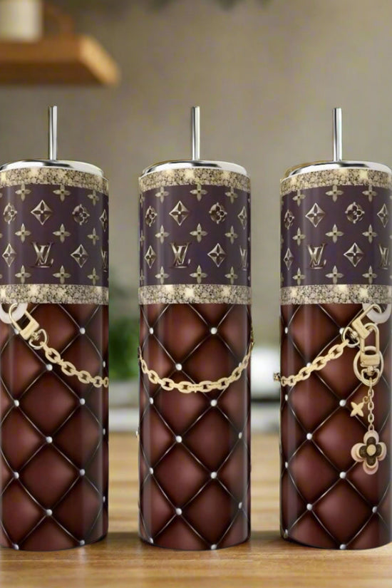 Three stylish reusable cups, featuring Kreative Kreationz's LV Brown Purse 20oz Tumbler with brown quilted patterns, gold chains, and monogram designs, are displayed on a wooden kitchen surface with a blurred background.