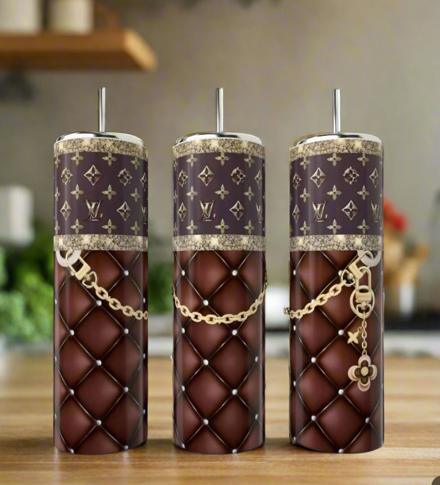 Three stylish reusable cups, featuring Kreative Kreationz's LV Brown Purse 20oz Tumbler with brown quilted patterns, gold chains, and monogram designs, are displayed on a wooden kitchen surface with a blurred background.