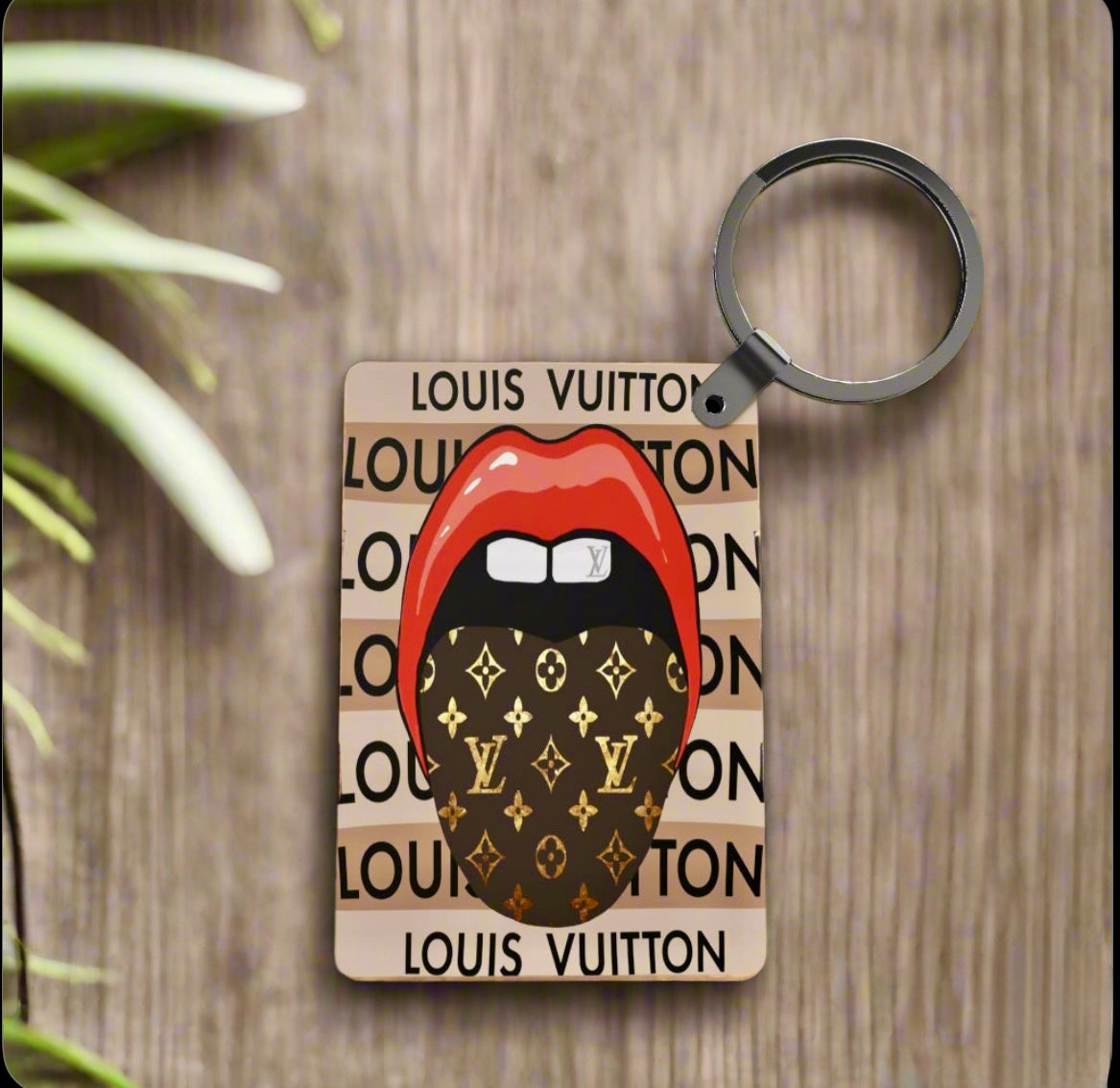 The LV Inspired Keychain by Kreative Kreationz features red lips and a tongue with Louis Vuitton pattern on a tan background with "LOUIS VUITTON" print. This stylish, lightweight keychain is shown resting on a wooden surface.