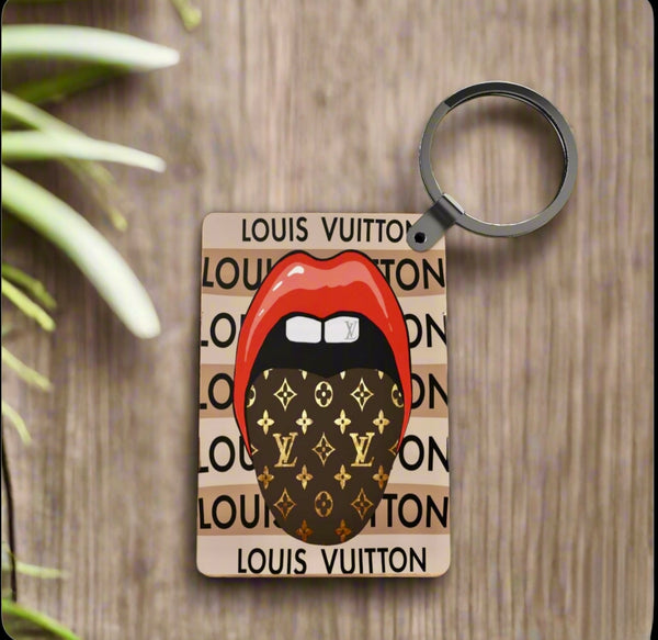The LV Inspired Keychain by Kreative Kreationz features red lips and a tongue with Louis Vuitton pattern on a tan background with "LOUIS VUITTON" print. This stylish, lightweight keychain is shown resting on a wooden surface.