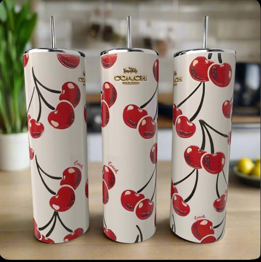Three white Kreative Kreationz Cherry Coach 20oz Skinny Tumblers with metal straws, featuring red cherry patterns and gold Coach-inspired logos, stand on a wooden kitchen surface—an eco-friendly, stylish choice.