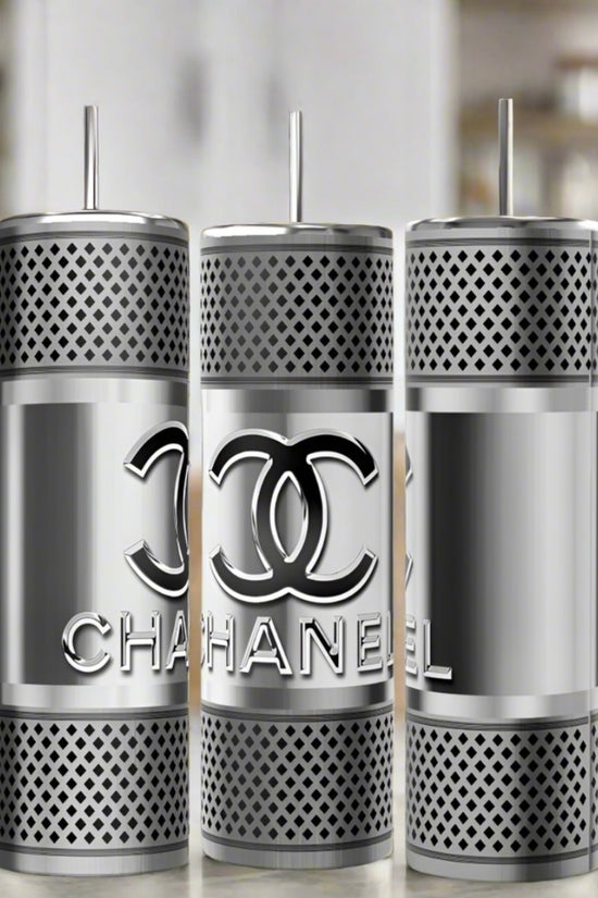 Three Chanel Silver Stainless Steel 20oz Tumblers by Kreative Kreationz are displayed with straws, showcasing a mesh pattern and bold overlapping C logo. Their shiny finish contrasts with the blurred modern kitchen backdrop.
