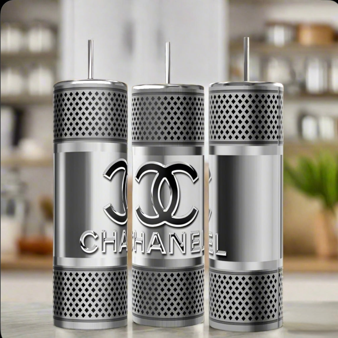 Three Chanel Silver Stainless Steel 20oz Tumblers by Kreative Kreationz are displayed with straws, showcasing a mesh pattern and bold overlapping C logo. Their shiny finish contrasts with the blurred modern kitchen backdrop.