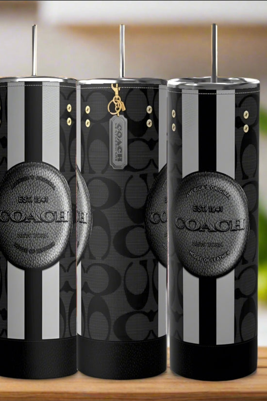 Three Kreative Kreationz Coach 20oz Tumblers featuring black and gray patterns, vertical stripes, Coach logos, metallic lids, and reusable straws are displayed on a wooden surface with a blurred background.