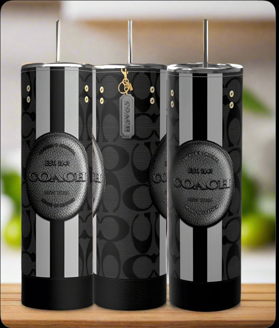 Three Kreative Kreationz Coach 20oz Tumblers featuring black and gray patterns, vertical stripes, Coach logos, metallic lids, and reusable straws are displayed on a wooden surface with a blurred background.