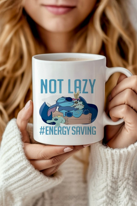 A woman in a white sweater holds the Stitch 15oz Mug by Kreative Kreationz, featuring a cartoon sloth lounging with the words “NOT LAZY #ENERGY SAVING” in a vibrant sublimation design close to her face.