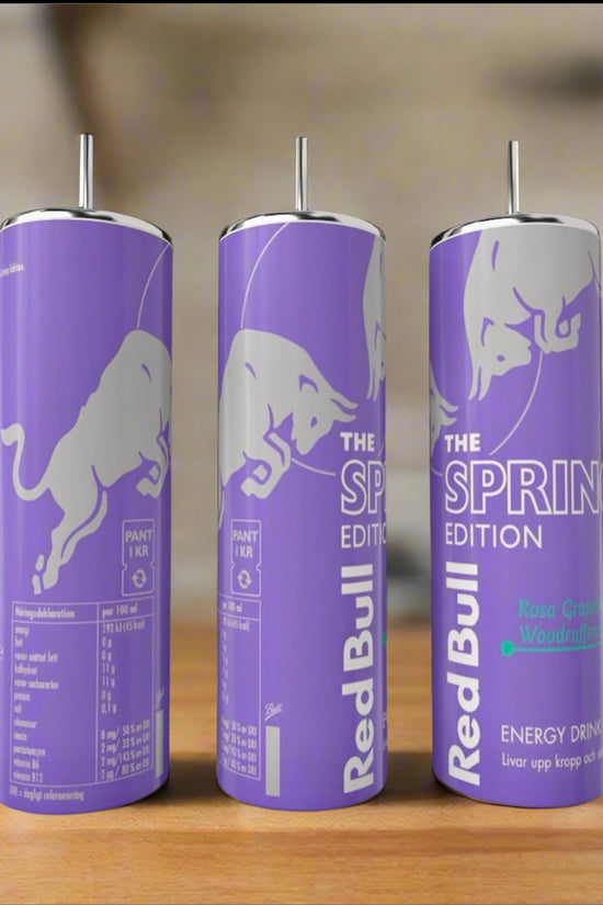 Three tall, lavender Kreative Kreationz Spring Red Bull 20oz Tumblers with silver bull graphics and eco-friendly reusable metal straws stand on a wooden surface, set against a blurred green background.