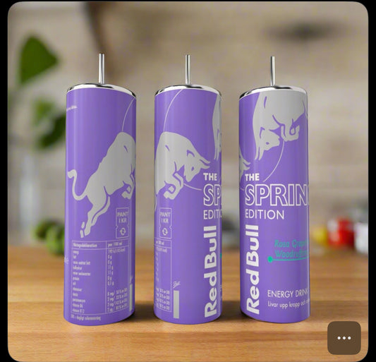 Three tall, lavender Kreative Kreationz Spring Red Bull 20oz Tumblers with silver bull graphics and eco-friendly reusable metal straws stand on a wooden surface, set against a blurred green background.