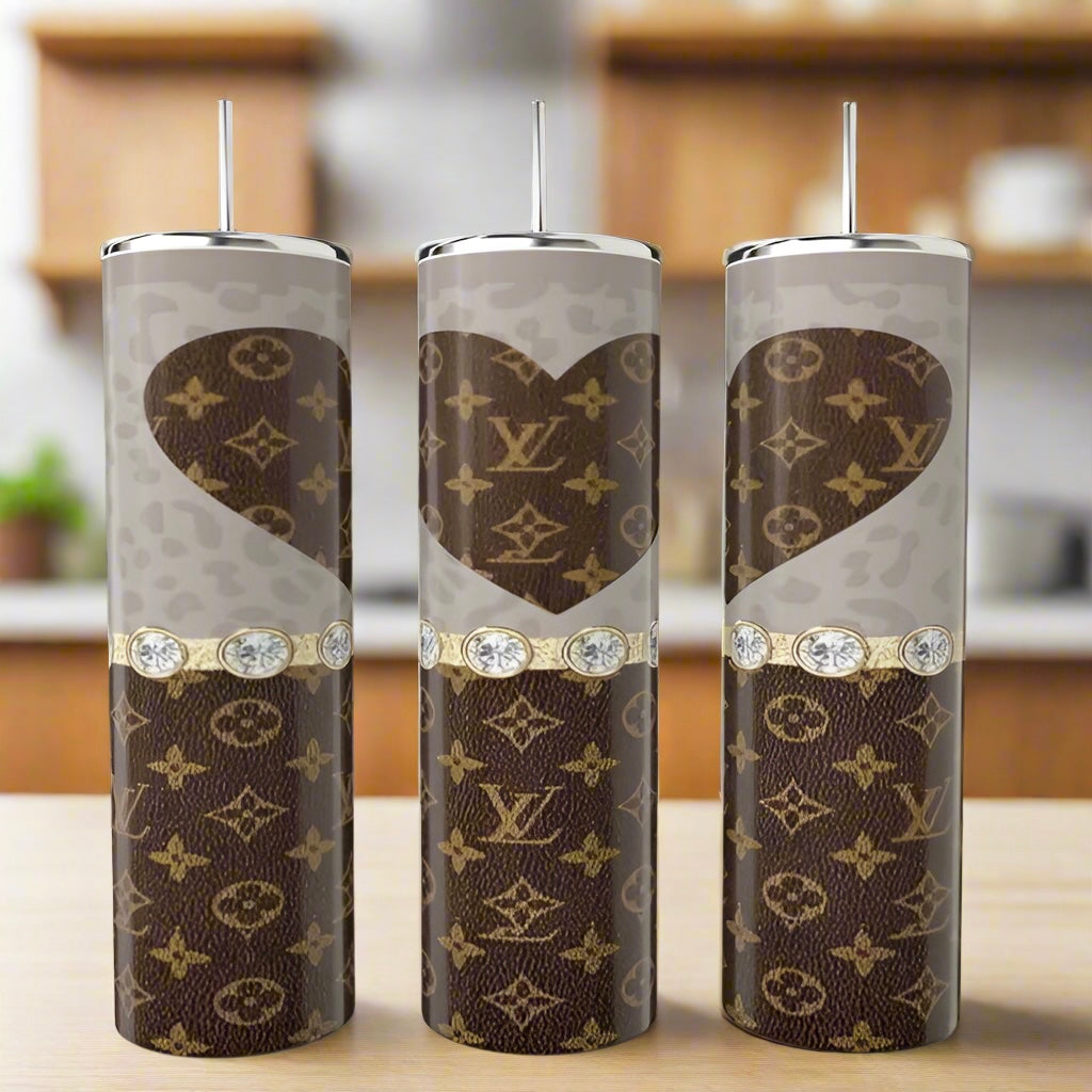 Three LV Heart 20oz Tumblers by Kreative Kreationz, with brown and beige Louis Vuitton-inspired patterns, gold accents, hearts, and diamond-like gems, are displayed on a kitchen counter.