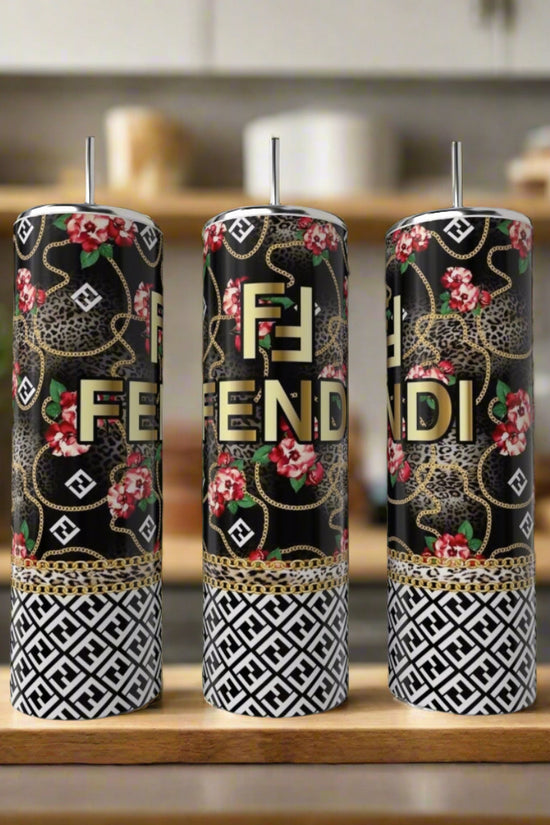 Three tall candles in black, gold, and white with Fendi-inspired designs (floral, leopard, geometric) sit beside a FENDI Rose 20oz Skinny Tumbler by Kreative Kreationz on a wooden surface in a modern kitchen.