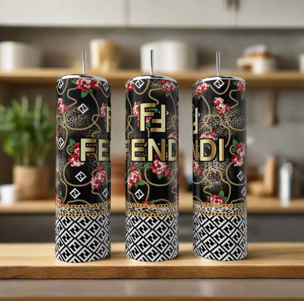 Three tall candles in black, gold, and white with Fendi-inspired designs (floral, leopard, geometric) sit beside a FENDI Rose 20oz Skinny Tumbler by Kreative Kreationz on a wooden surface in a modern kitchen.