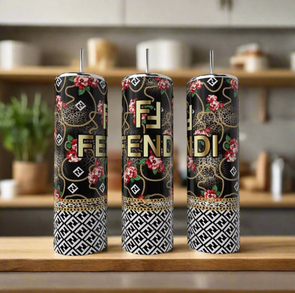 Three tall candles in black, gold, and white with Fendi-inspired designs (floral, leopard, geometric) sit beside a FENDI Rose 20oz Skinny Tumbler by Kreative Kreationz on a wooden surface in a modern kitchen.
