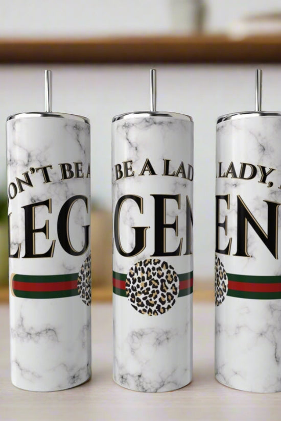 Three white marble “Don’t Be a Lady Be a LEGEND” Gucci 20oz Skinny Tumblers by Kreative Kreationz, each with bold black text, green and red stripe, leopard print circle, and metal straws, are displayed on a table amid blurred shelves and plants.