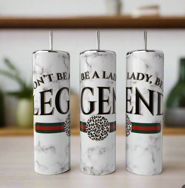 Three white marble “Don’t Be a Lady Be a LEGEND” Gucci 20oz Skinny Tumblers by Kreative Kreationz, each with bold black text, green and red stripe, leopard print circle, and metal straws, are displayed on a table amid blurred shelves and plants.