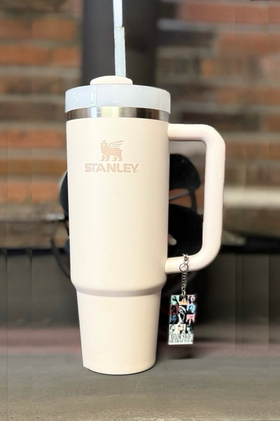 A light pink Stanley tumbler with a straw sits on a wooden surface. Hanging from the handle is Kreative Kreationz’s ERAS Charm Dangle, a handcrafted cup accessory featuring photos and charms. A blurred brick wall is in the background.