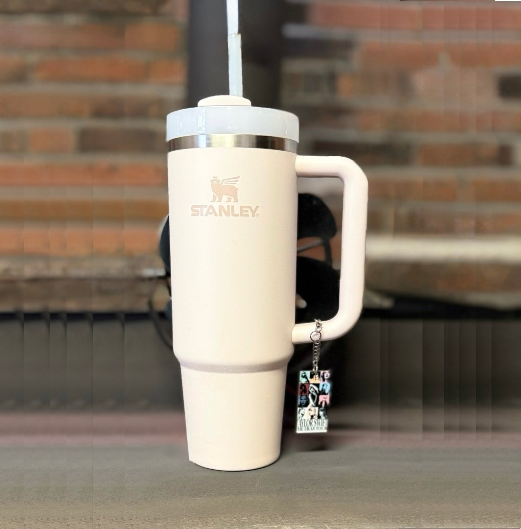 A light pink Stanley tumbler with a straw sits on a wooden surface. Hanging from the handle is Kreative Kreationz’s ERAS Charm Dangle, a handcrafted cup accessory featuring photos and charms. A blurred brick wall is in the background.