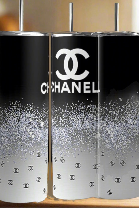 Three Chanel | Black and Glitter 20oz Skinny Tumblers by Kreative Kreationz sit on wood. Each features a sleek black-to-white gradient, glittery specks, and the Chanel logo, creating an elegant look against a blurred kitchen backdrop.
