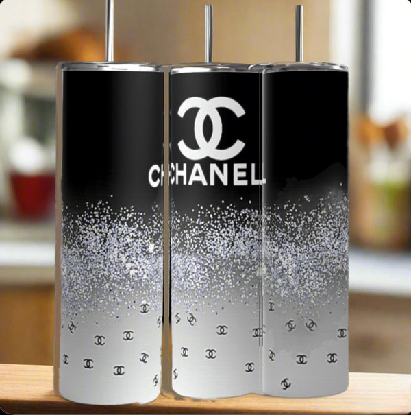 Three Chanel | Black and Glitter 20oz Skinny Tumblers by Kreative Kreationz sit on wood. Each features a sleek black-to-white gradient, glittery specks, and the Chanel logo, creating an elegant look against a blurred kitchen backdrop.