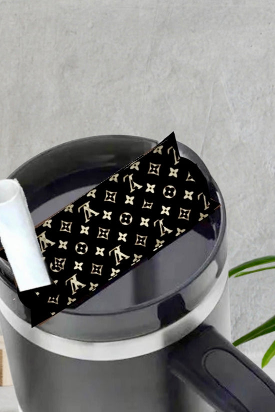 The LV | Stanley Lid Plate/Topper by Kreative Kreationz sits atop a black coffee mug with a white straw, featuring a gold and black designer-style pattern. Behind it, a green plant and wooden block rest on a gray surface.