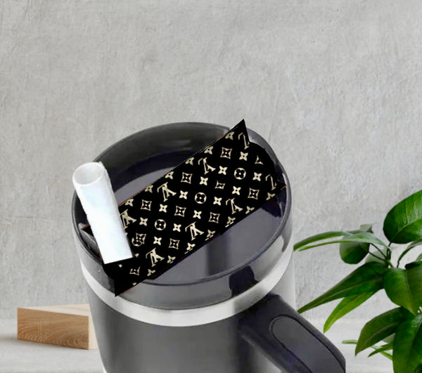 The LV | Stanley Lid Plate/Topper by Kreative Kreationz sits atop a black coffee mug with a white straw, featuring a gold and black designer-style pattern. Behind it, a green plant and wooden block rest on a gray surface.