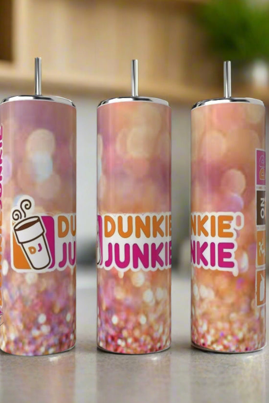 Three Kreative Kreationz Dunkin Junkie 20oz Tumblers with reusable straws sit on a counter. Each features a pink, orange, and white gradient design, “DUNKIN JUNKIE” text, and a coffee cup graphic for a fun touch.