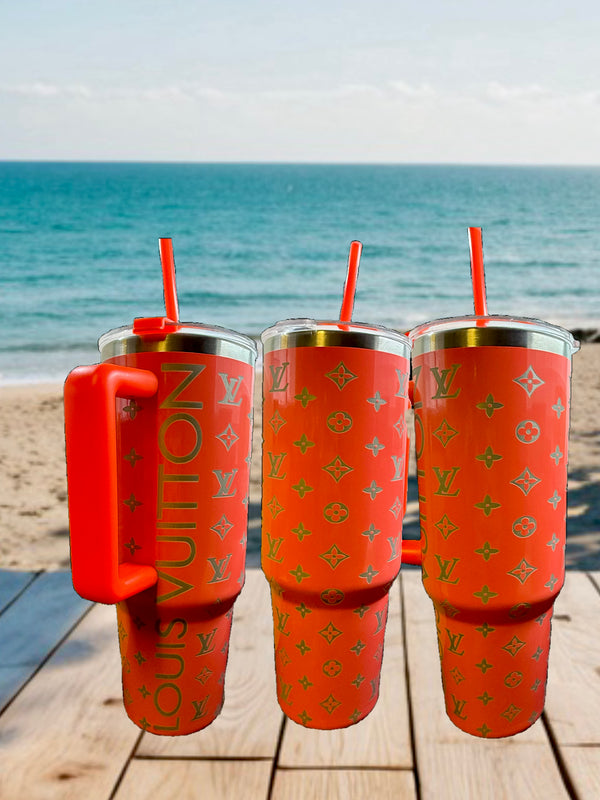Three Kreative Kreationz Coral 40oz LV Engraved Tumblers with straws rest on a wooden deck, overlooking a sandy beach and the ocean beneath a bright blue sky.