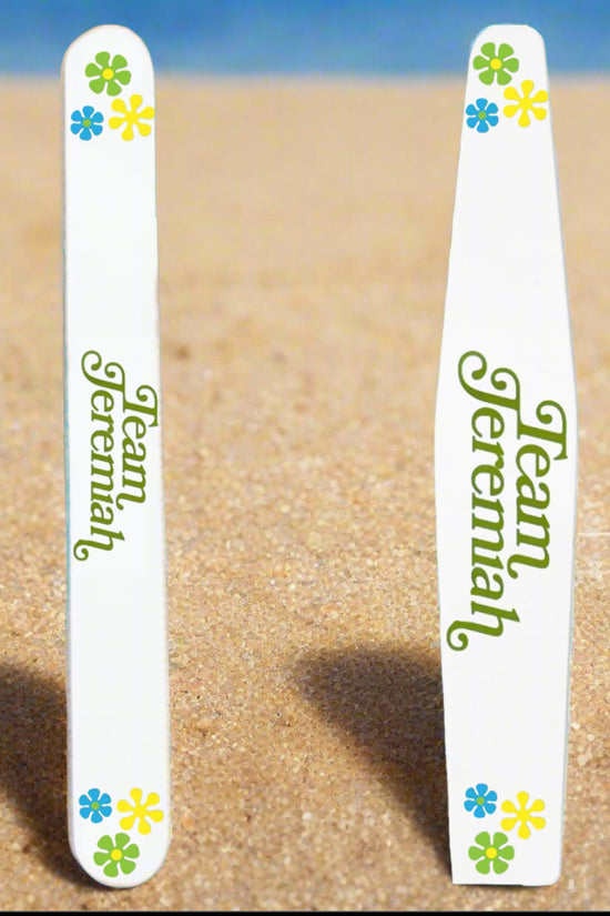 Two Kreative Kreationz Team Jeremiah Ceramic Nail Files featuring green text and colorful flowers stand upright on sandy beach, with a blurred blue sky or water in the background.