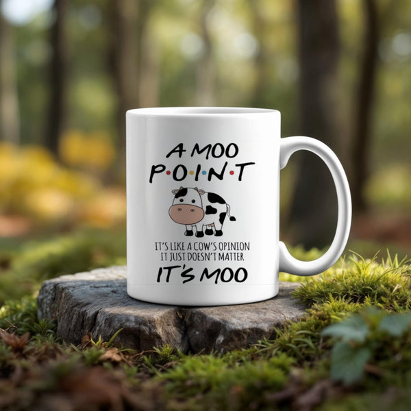 The Friends Moo Point Mug by Kreative Kreationz—a white ceramic mug with a cartoon cow and the quote, "A MOO POINT. It's like a cow's opinion, it just doesn't matter. It's moo"—rests on a mossy tree stump in a sunlit forest.
