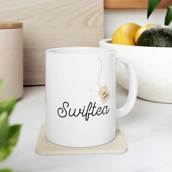 A Kreative Kreationz Swiftea Mug est 1989, featuring "Swiftea" and a tea bag tag icon with "Est. 1989," gracefully sits on a marble counter beside lemons and an avocado, highlighting its original artwork.