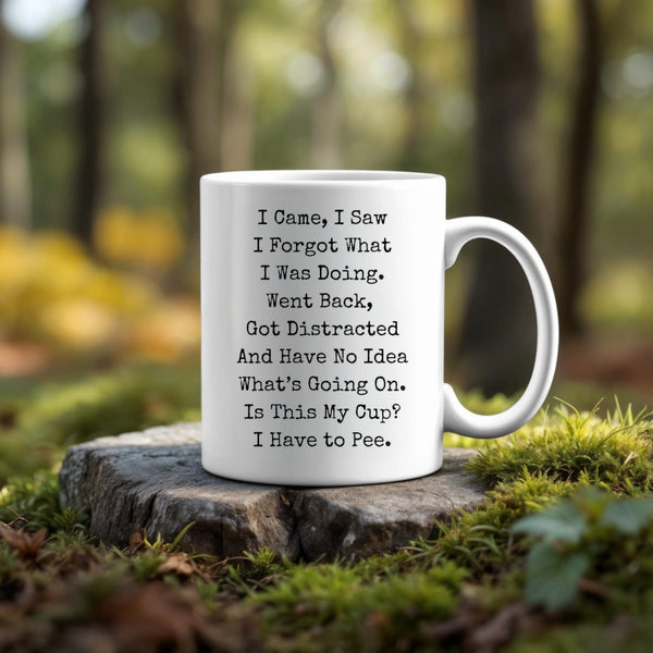 The ADHD Ceramic Mug by Kreative Kreationz, a white dishwasher-safe mug with funny black text—"I came, I saw I forgot what I was doing. Went back, got distracted and have no idea what's going on."—rests on a tree stump in a sunlit forest.