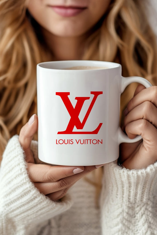A woman in a cozy white sweater holds the LV Ceramic Mug by Kreative Kreationz, featuring a red Louis Vuitton-style logo, and smiles as she enjoys her drink.