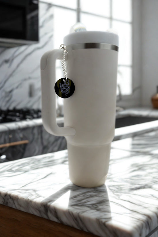 A large white insulated mug with a handle rests on a marble countertop in sunlight. Hanging from its lid is the Kreative Kreationz REP|13 Snake Engraved Cup Charm featuring a coiled snake and 