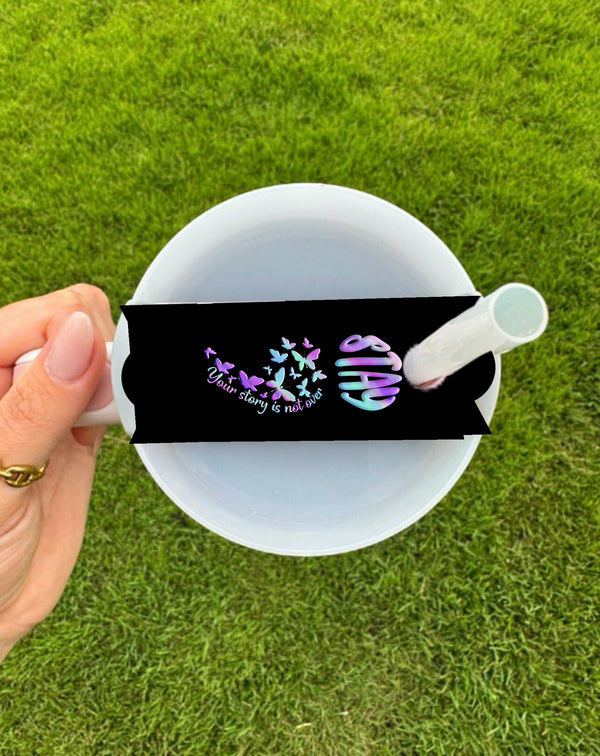 A hand holds a customizable STAY Suicide Awareness Stanley Lid Topper by Kreative Kreationz, featuring a black band with colorful butterflies and the phrase "Your story is not over," along with a rainbow-colored semicolon. The grassy backdrop enhances this whimsical design.
