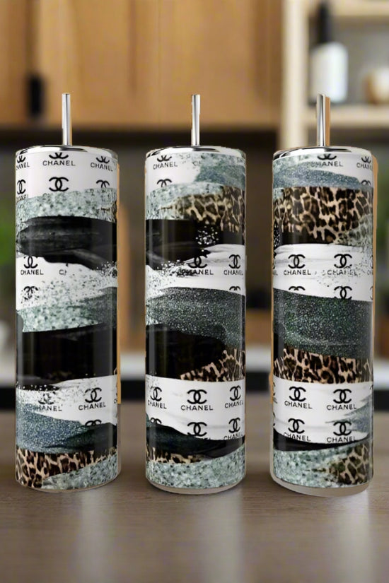 Three Kreative Kreationz 20oz stainless steel tumbler cups, including a leopard print, Chanel Stripe Design, and a sleek black-gray style, rest on wood with metal straws. Their durability shines against a blurred kitchen backdrop with potted plants.