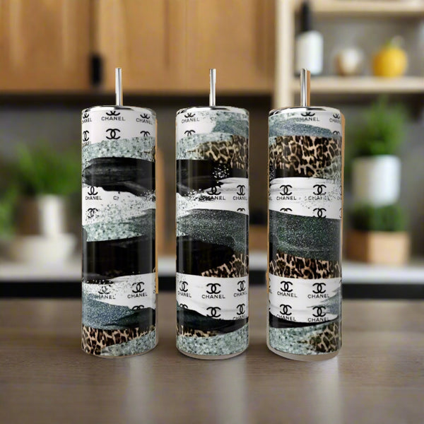 Three Kreative Kreationz 20oz stainless steel tumbler cups, including a leopard print, Chanel Stripe Design, and a sleek black-gray style, rest on wood with metal straws. Their durability shines against a blurred kitchen backdrop with potted plants.
