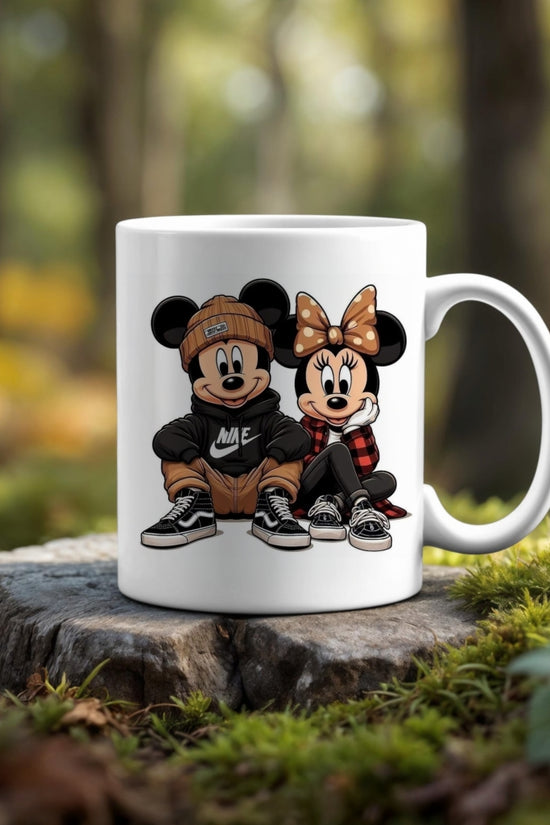 The 11oz Ceramic Mug by Kreative Kreationz showcases cartoon characters styled as Mickey and Minnie Mouse in trendy streetwear, sitting side by side. This dishwasher-safe mug is pictured outdoors on a tree stump with a blurred forest background.