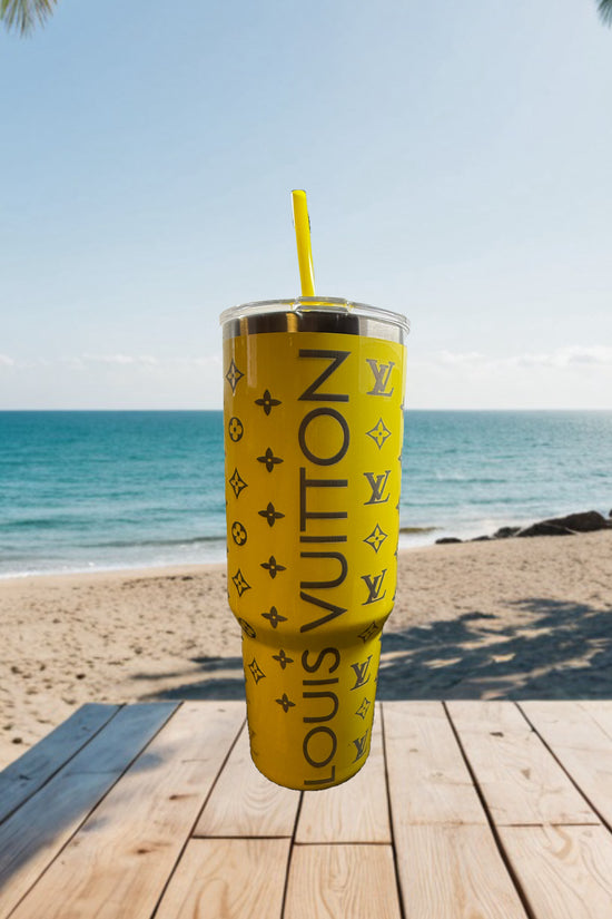 A yellow LV Engraved Tumbler by Kreative Kreationz with a straw sits on a wooden table by the sandy beach, ocean, and clear sky in the background.