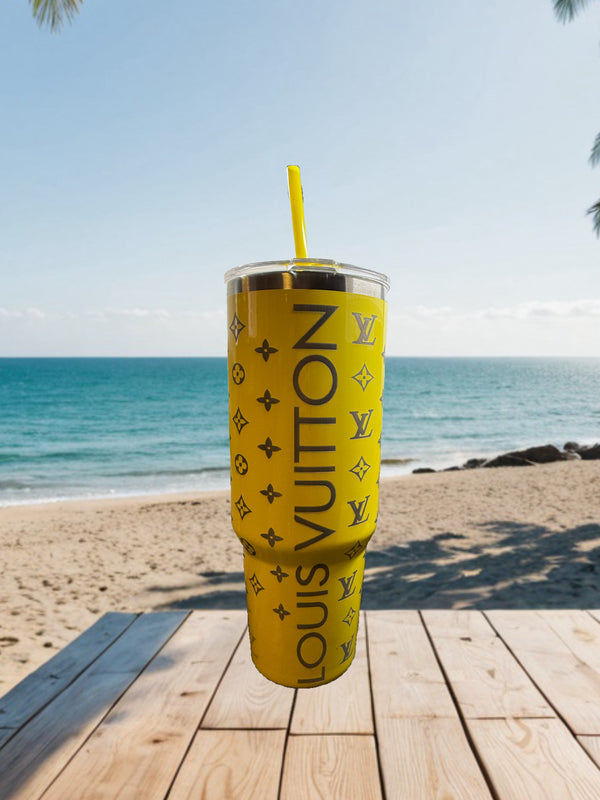A yellow LV Engraved Tumbler by Kreative Kreationz with a straw sits on a wooden table by the sandy beach, ocean, and clear sky in the background.
