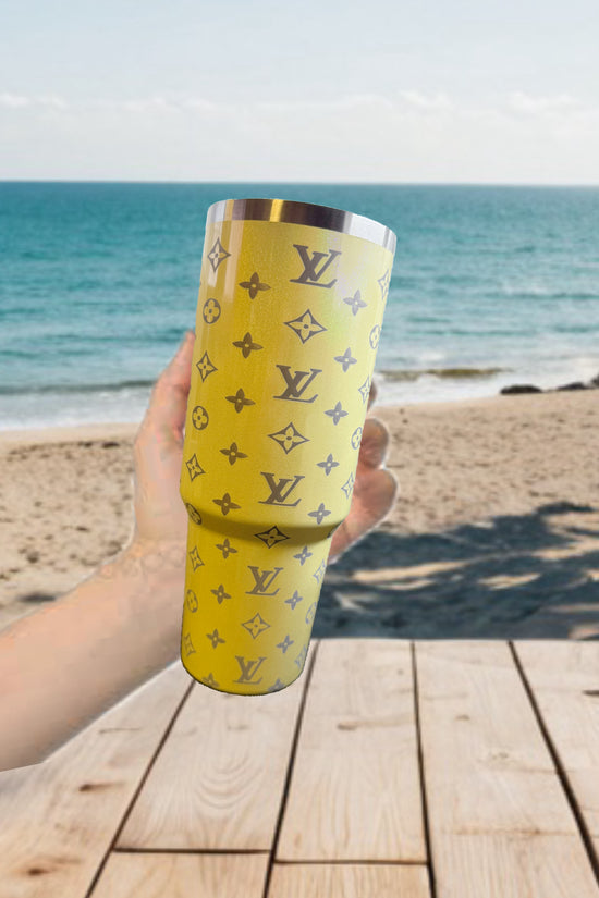 A hand holds the Yellow Shimmer 40oz LV Engraved Tumbler by Kreative Kreationz, featuring a Louis Vuitton-style pattern, with a sandy beach, ocean waves, and a wooden table in the background.