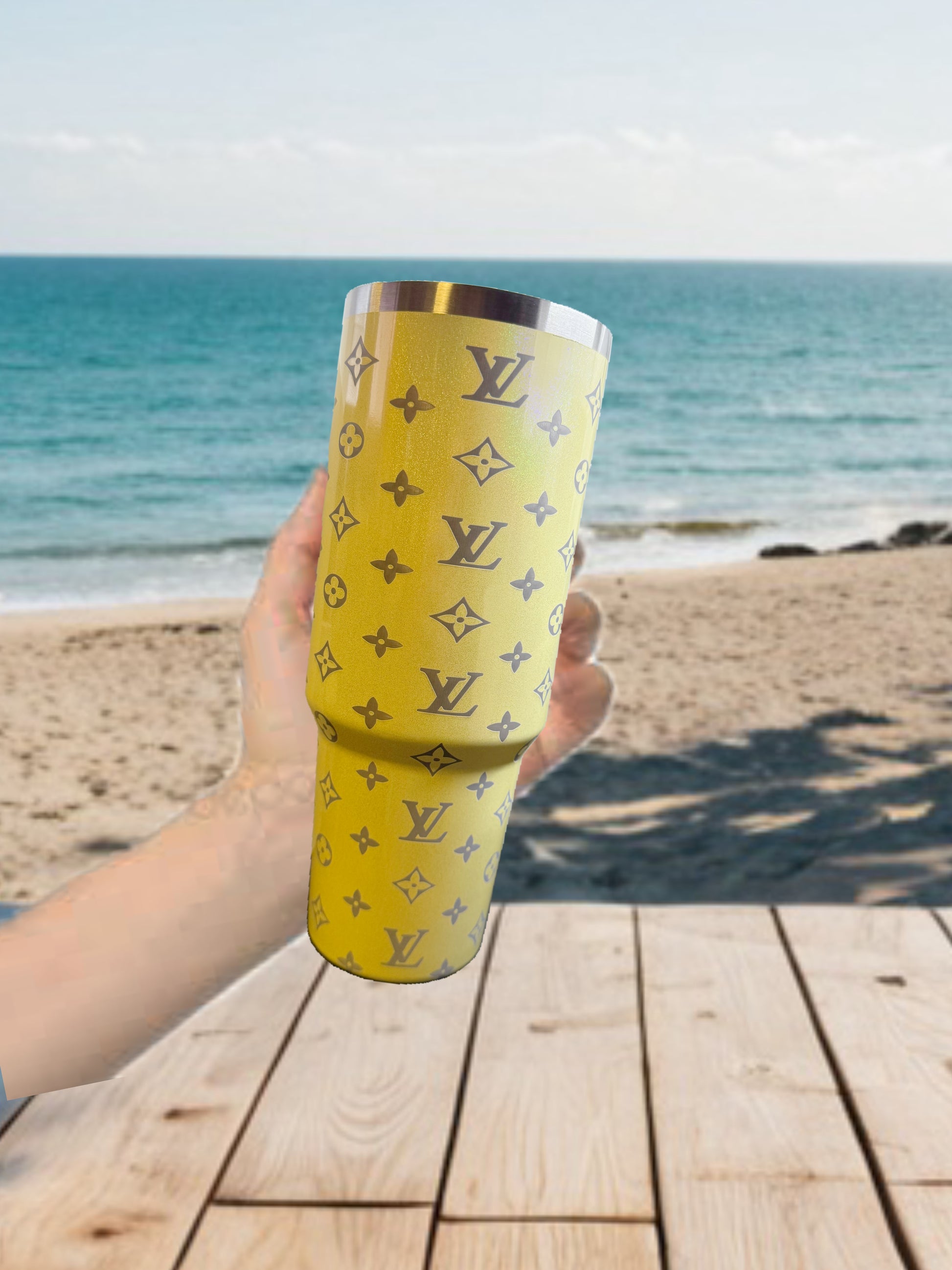 A hand holds the Yellow Shimmer 40oz LV Engraved Tumbler by Kreative Kreationz, featuring a Louis Vuitton-style pattern, with a sandy beach, ocean waves, and a wooden table in the background.