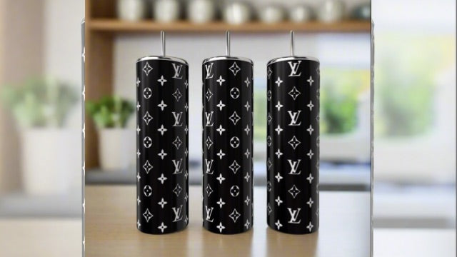 Three Black & White LV 20oz Tumblers by Kreative Kreationz, each featuring a white monogram on black stainless steel and metal straws, are displayed on a table with a blurred kitchen backdrop, highlighting their luxury-inspired style.