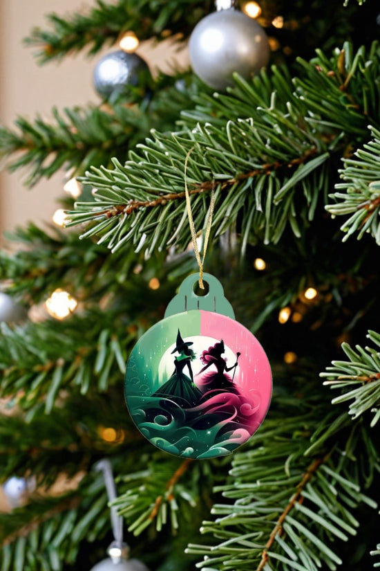 A close-up of a decorated Christmas tree highlights Kreative Kreationz's Wicked Christmas Tree Ornament, showcasing a whimsical illustration of a witch and a girl in a magical forest. The tree is adorned with silver baubles and pine needles, making it ideal for festive decorators seeking enchanting holiday decor.
