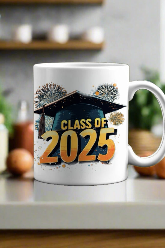 A Kreative Kreationz Class of 2025 Graduate Mug sits on a kitchen counter, featuring a graduation cap and fireworks. Blurry kitchen items are visible in the background. Satisfaction Guaranteed with every purchase.