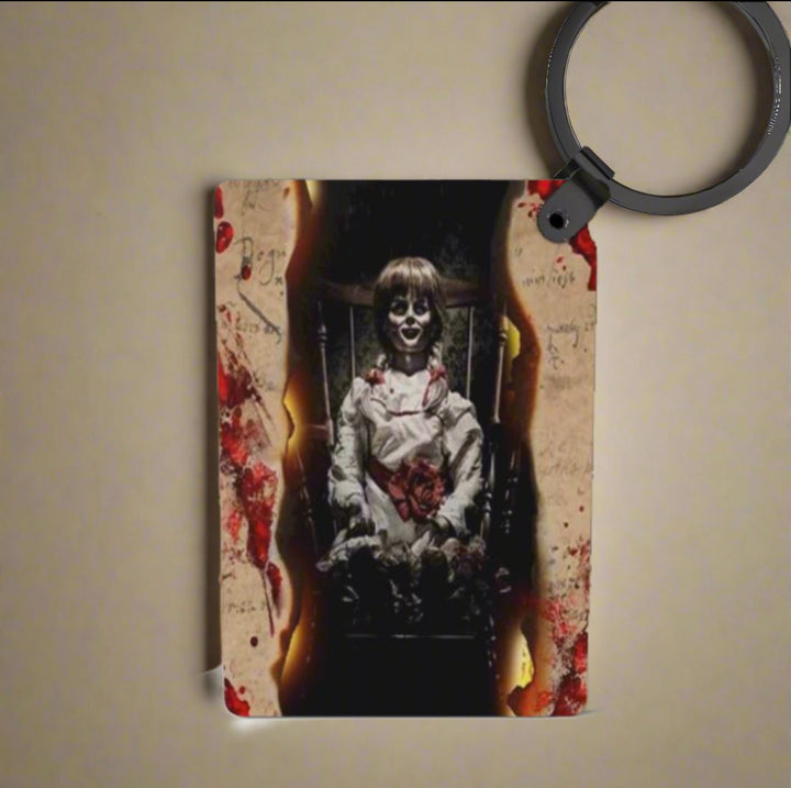 The Annabelle Inspired Keychain by Kreative Kreationz features a haunting porcelain doll with dark eyes and a creepy smile, set against burnt, bloodstained paper—a unique, personalized gift.