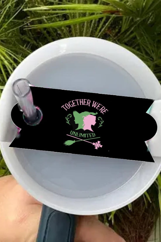 A hand holds a white tumbler with a straw and the Wicked -Stanley Lid Topper by Kreative Kreationz. A black sticker features a broom, wand, “TOGETHER WE’RE UNLIMITED,” and two face silhouettes. Green palm leaves are in the background.