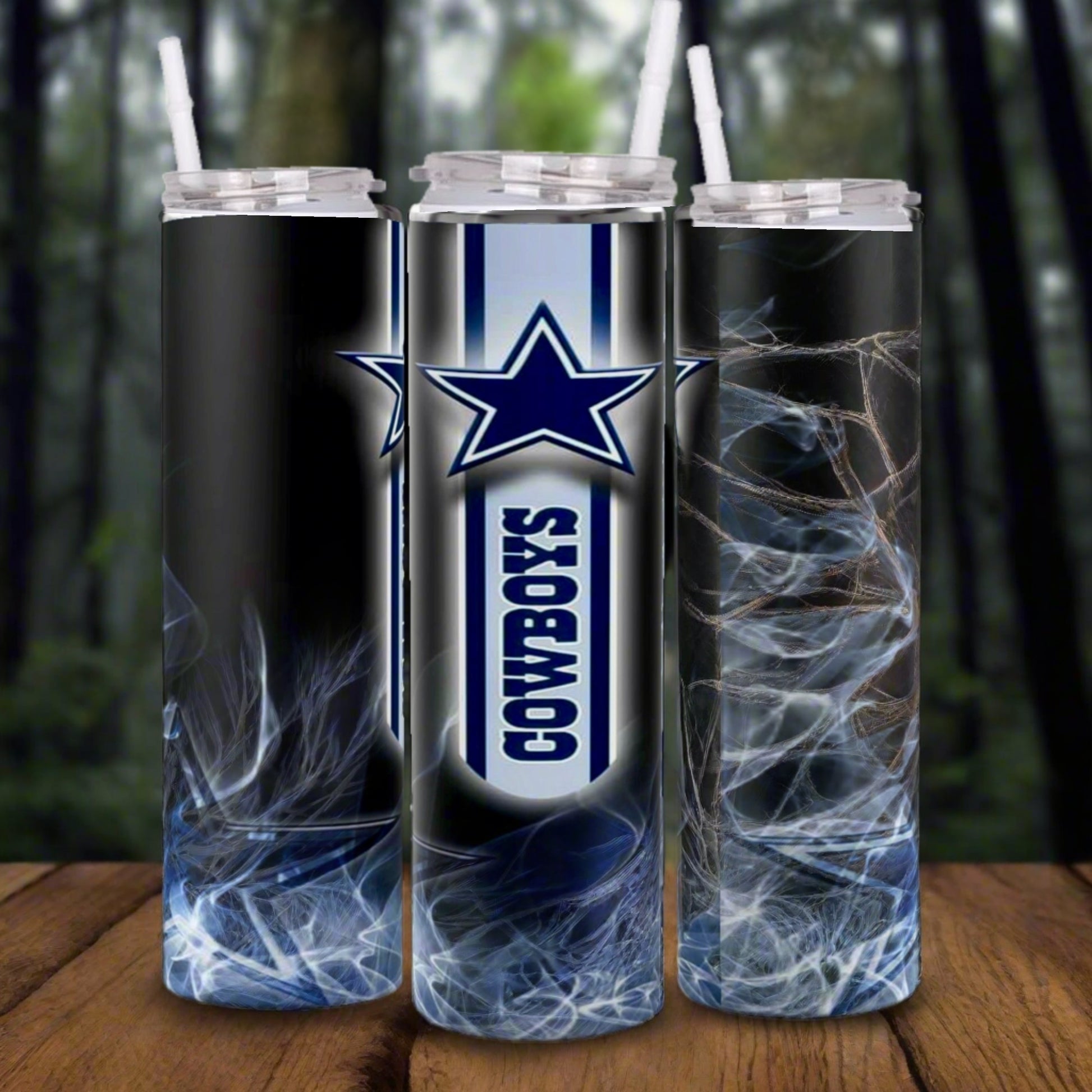 Three Kreative Kreationz Dallas Cowboys NFL Custom 20oz Skinny Tumblers with clear lids and straws, featuring the team's star logo and smoky abstract patterns, are displayed on a wooden surface with a blurred forest background.