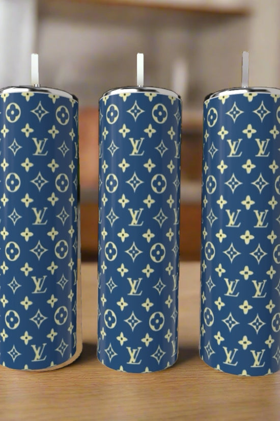 Three cylindrical candles with blue exteriors and white Louis Vuitton monogram patterns stand upright on a wooden surface, paired with a Kreative Kreationz LV Blue & Yellow 20oz Tumbler in the softly blurred indoor background.