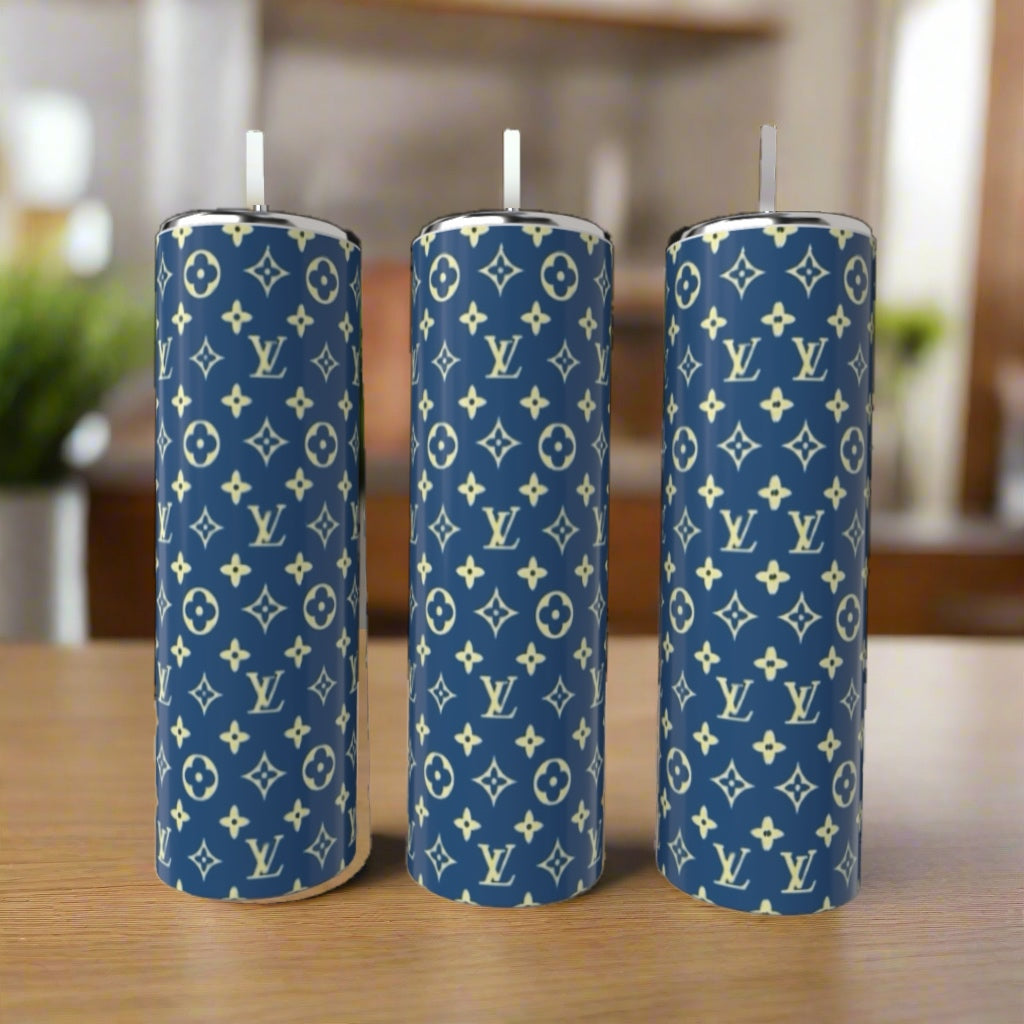 Three cylindrical candles with blue exteriors and white Louis Vuitton monogram patterns stand upright on a wooden surface, paired with a Kreative Kreationz LV Blue & Yellow 20oz Tumbler in the softly blurred indoor background.